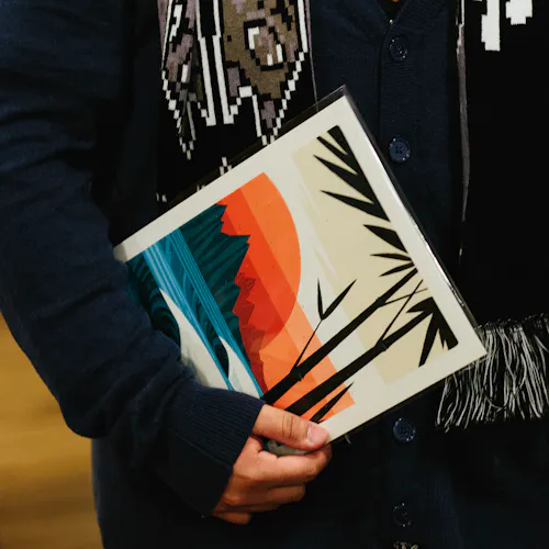 A man wearing a navy blue jacket and black scarf holding an art print wrapped in cellophane by his side.