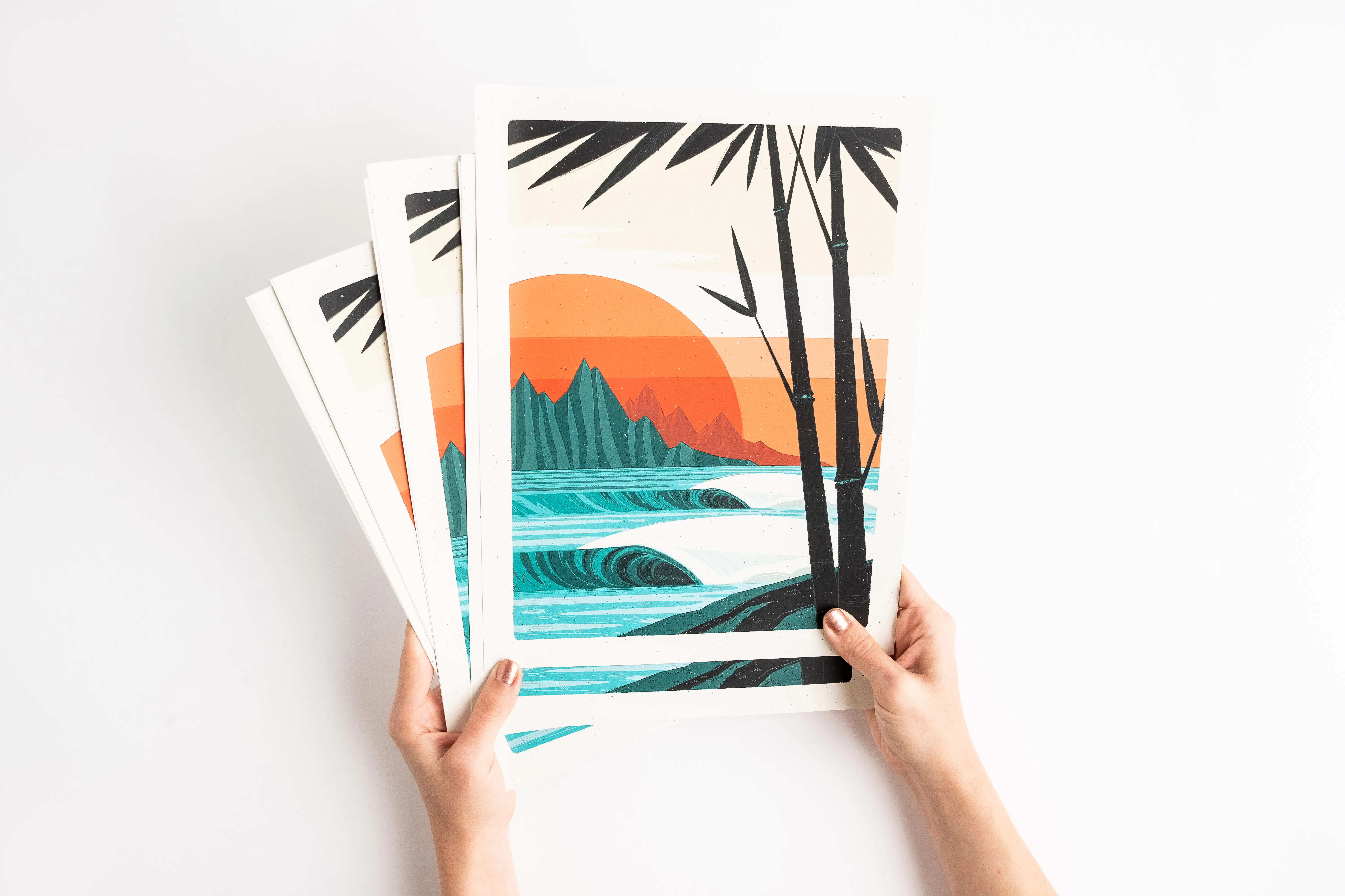 Two hands holding fanned-out art prints with a beach sunset design in shades of orange, blue and brown.
