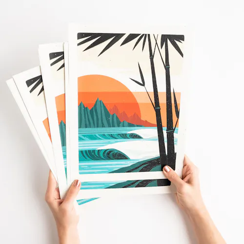 Two hands holding fanned-out art prints with a beach sunset design in shades of orange, blue and brown.