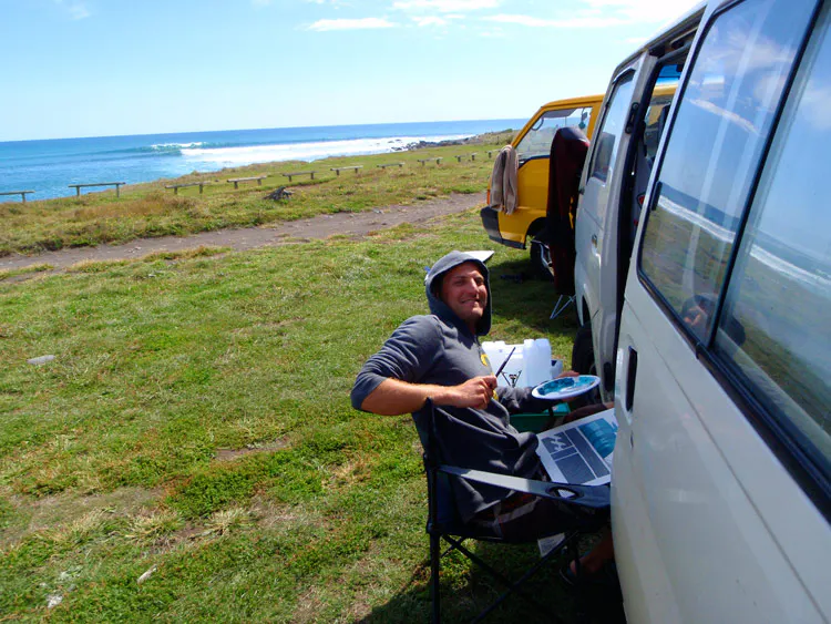 A man sitting and painting next to a white van parked in a grassy area with the ocean in front of it.