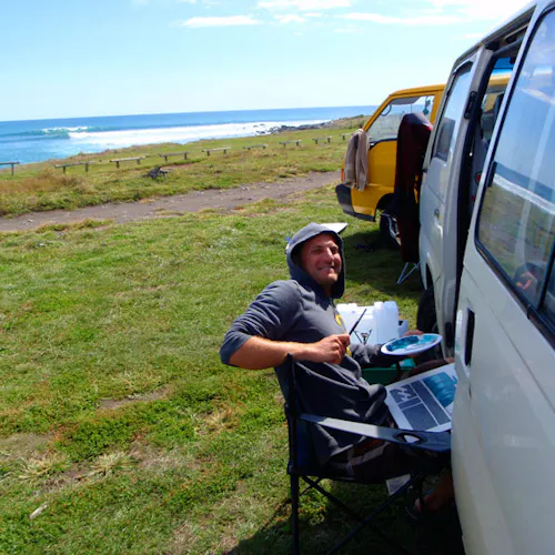 A man sitting and painting next to a white van parked in a grassy area with the ocean in front of it.