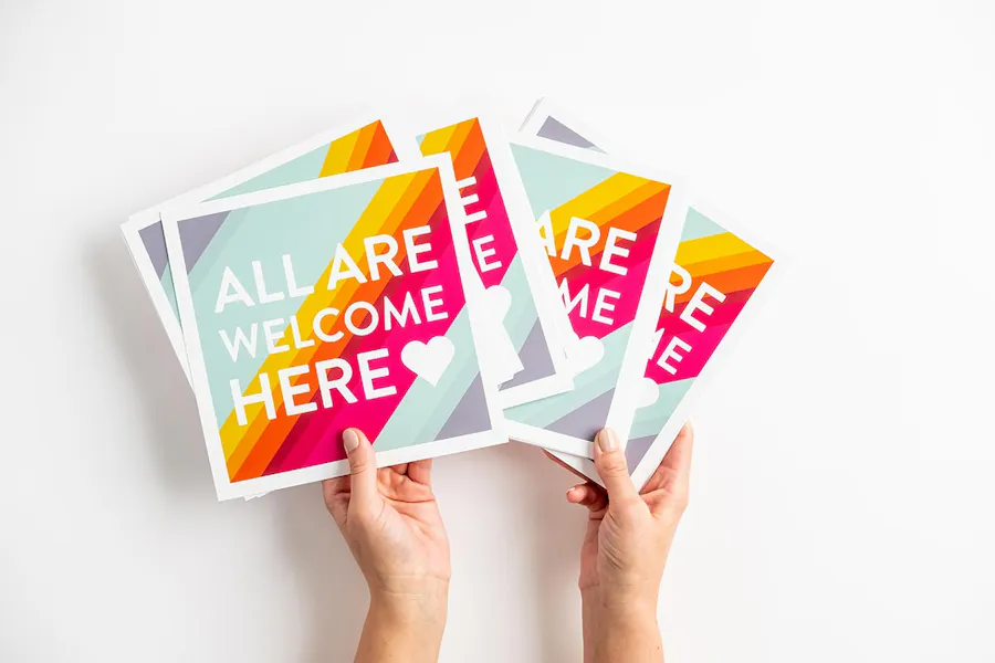 Two hands holding fanned-out signs with All are Welcome Here in white letters.