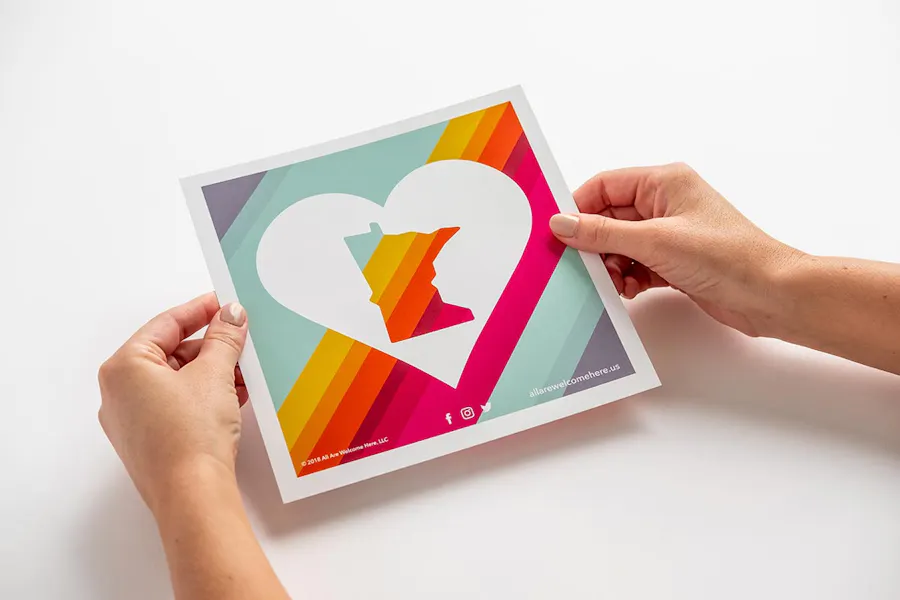 Two hands holding a sign with an outline of Minnesota inside a white heart against a bright striped background.