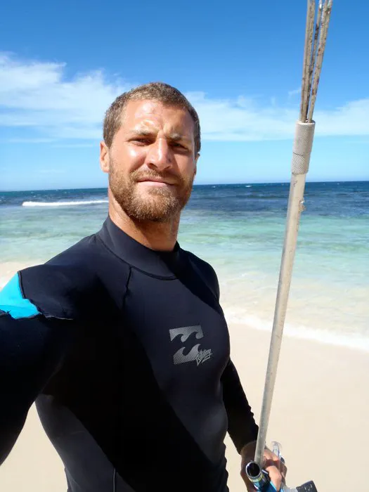 A man with a beard standing on the beach wearing a black wetsuit and holding a metal staff.