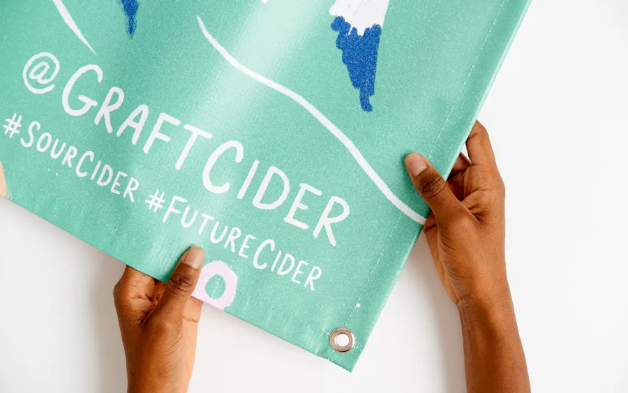 Two hands holding the corner of a custom printed banner with a grommet and Graft Cider in white letters.