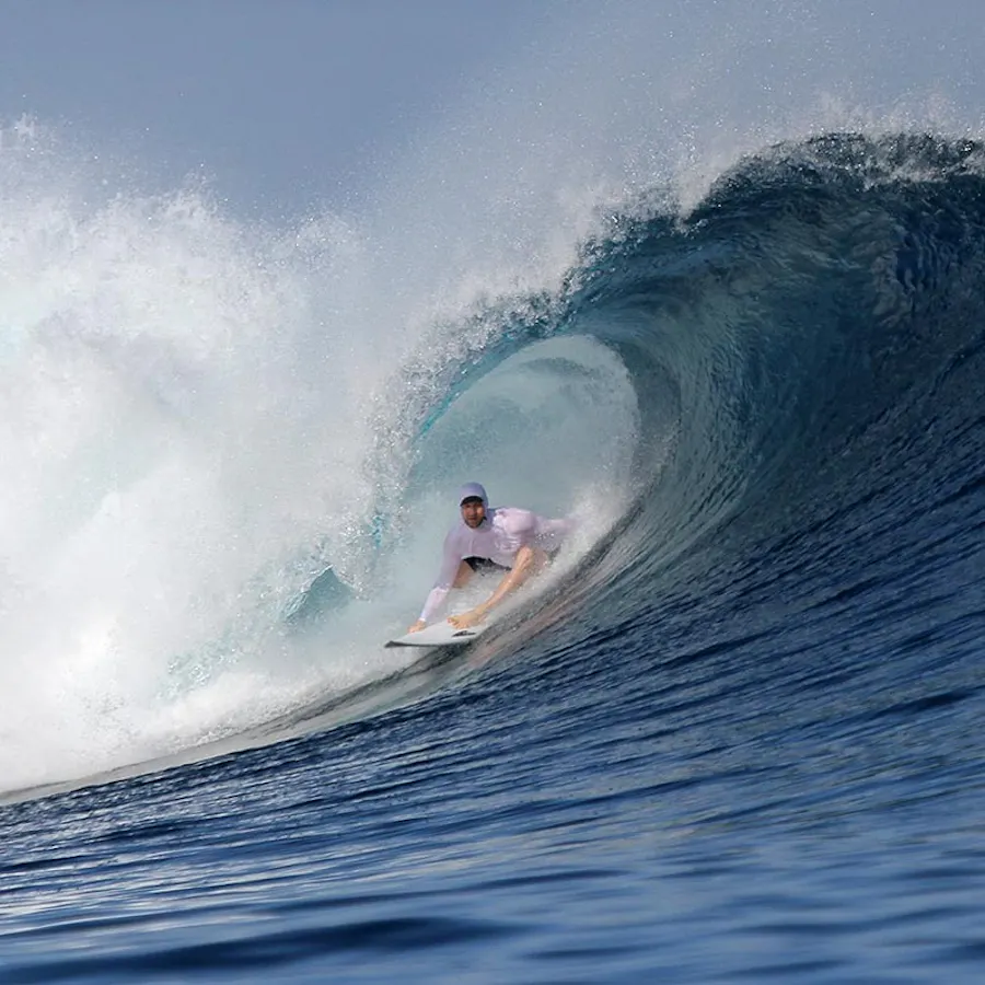 A man surfing in the ocean wearing a white hat, white top and black shorts.