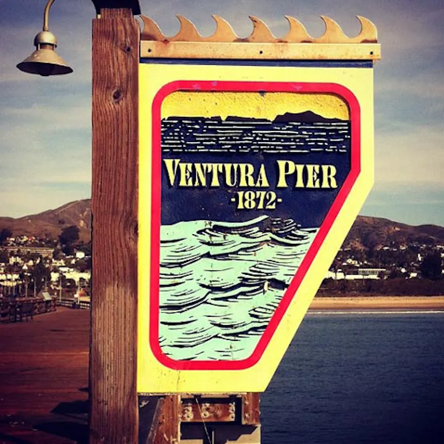 A sign on a pier lamp post with Ventura Pier 1872 in yellow and a teal waves design below it.