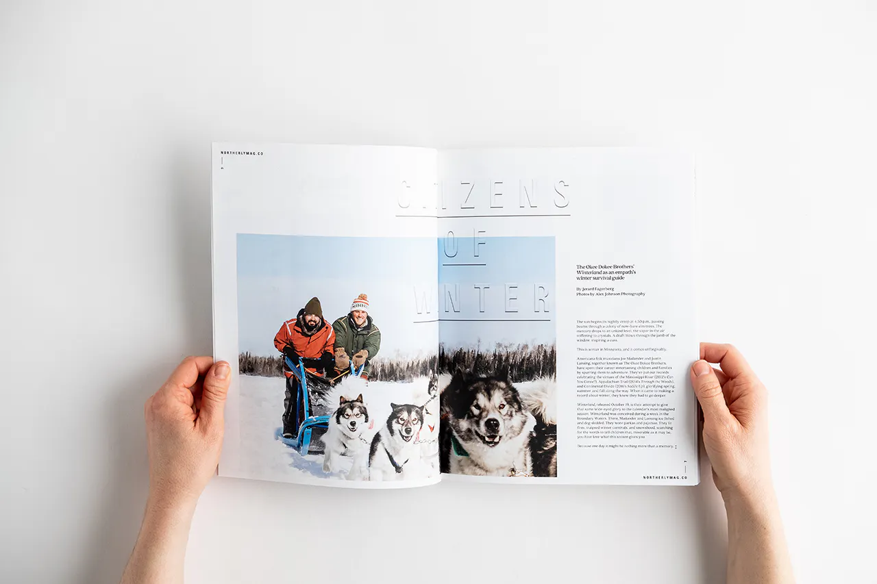 Two hands holding a photography magazine open to images of two people dogsledding in snow.
