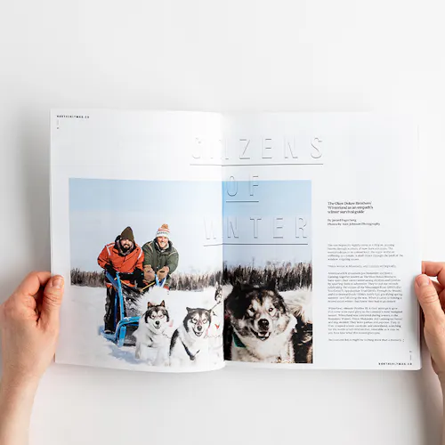 Two hands holding a photography magazine open to images of two people dogsledding in snow.