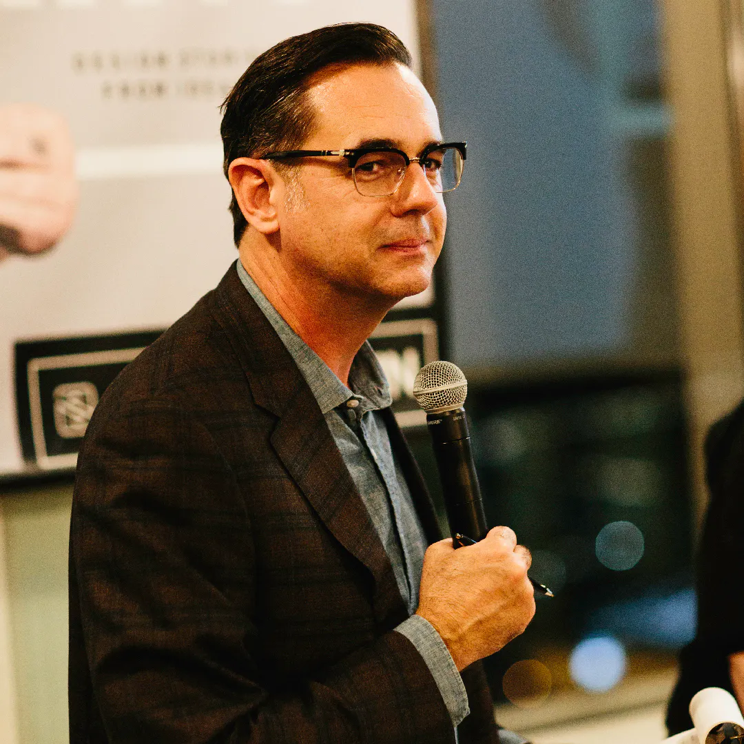 A man wearing glasses sitting on a stool and holding a microphone during a presentation.