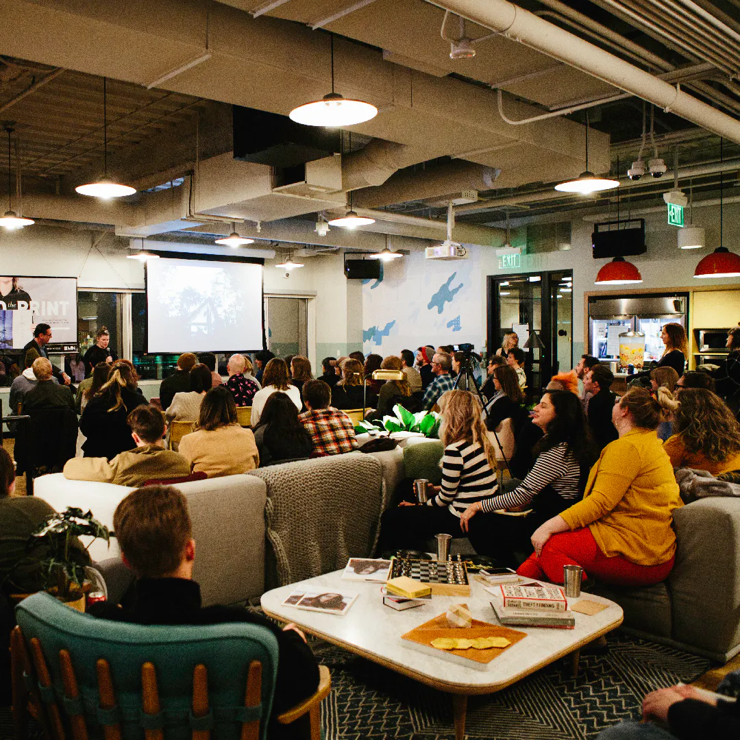 Two people standing in front a custom hanging banners with an audience sitting on couches and chairs.