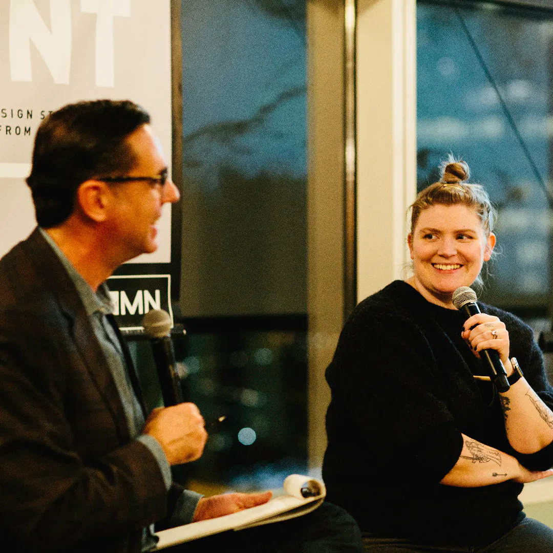 A man and a woman sitting on stools and holding microphones during a presentation.