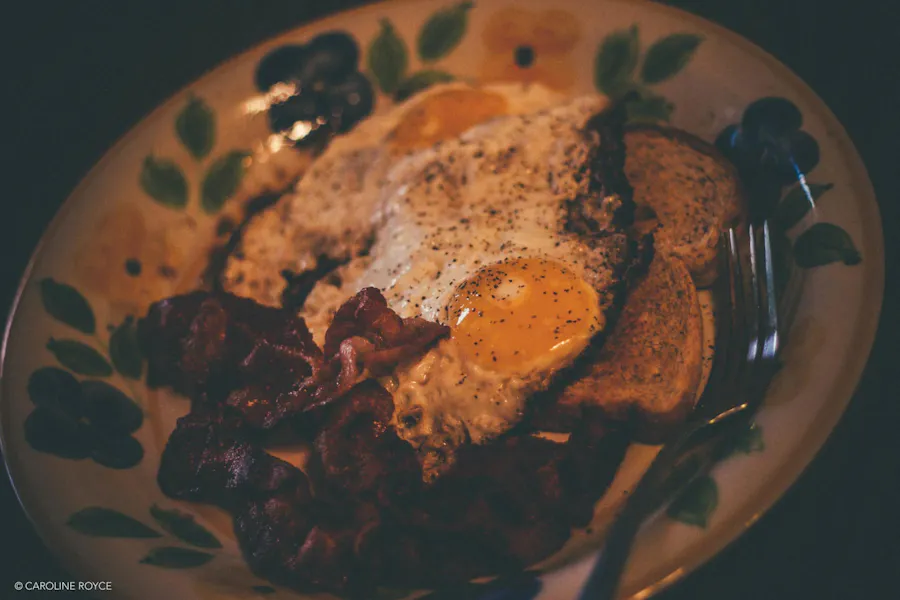 A dinner plate with a floral design along the edge and bacon, two eggs, toast and a fork on it.