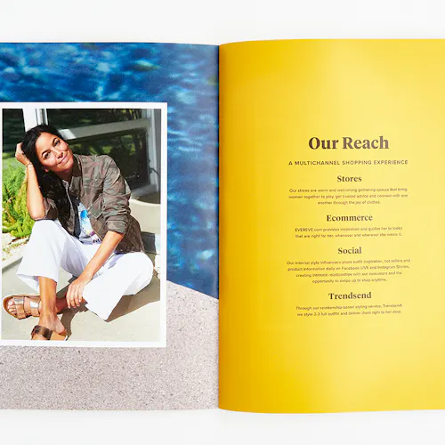 An Evereve brand book laying open to a woman sitting outside in a camo jacket and white pants and Our Reach in black.