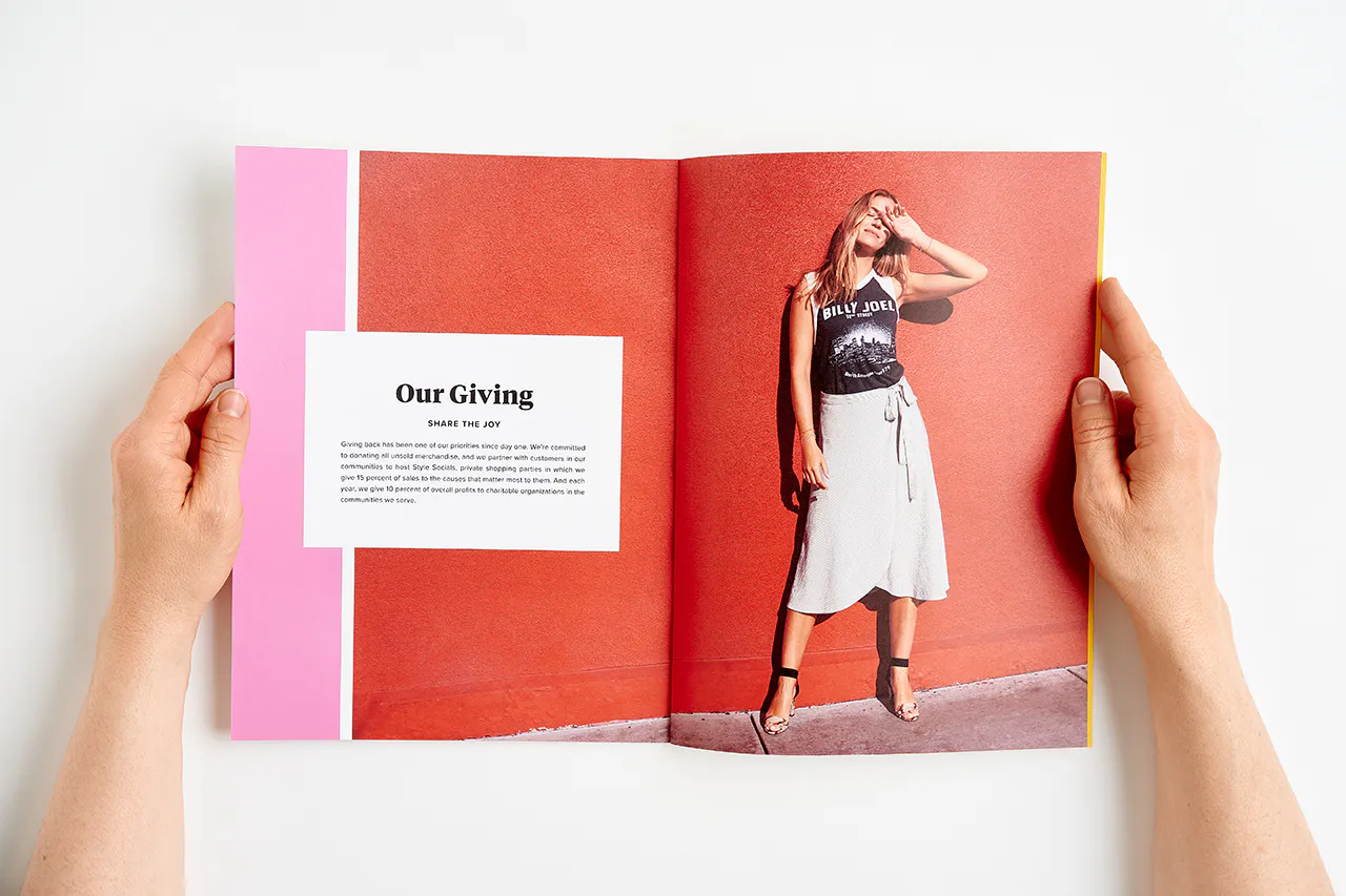 Two hands holding open an Evereve brand book to a woman in a white skirt standing against a red wall and Our Giving in black.
