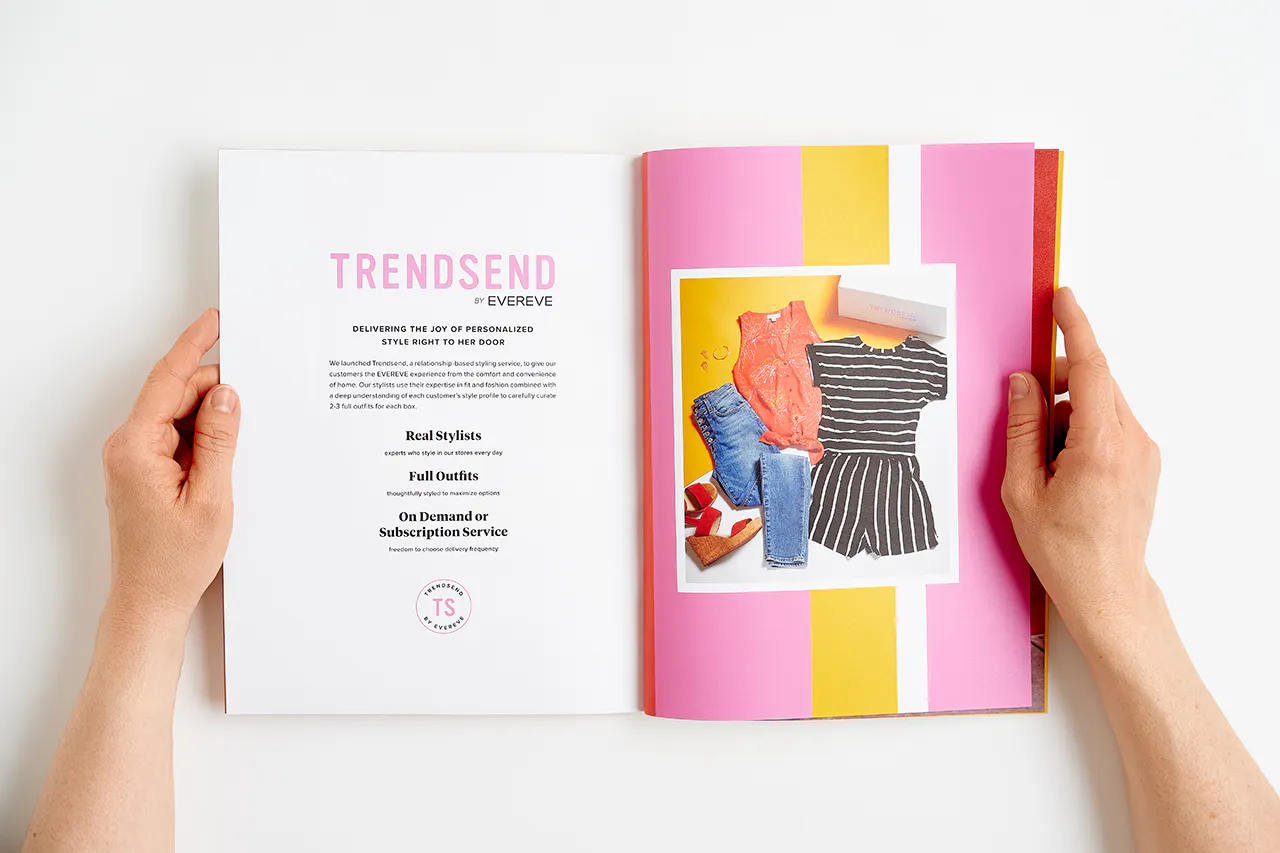 Two hands holding open an Evereve brand book to images of various clothing and Trendsend by Evereve.
