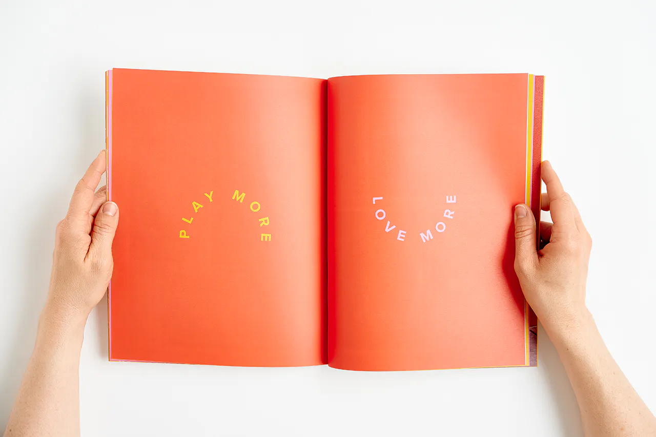 Two hands holding open an Evereve brand book to Play More and Love More on an orange background.