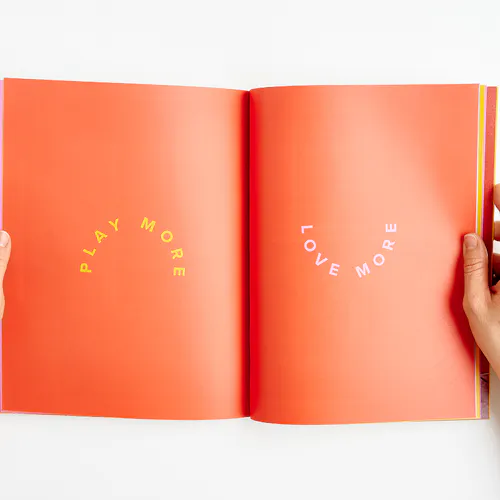 Two hands holding open an Evereve brand book to Play More and Love More on an orange background.