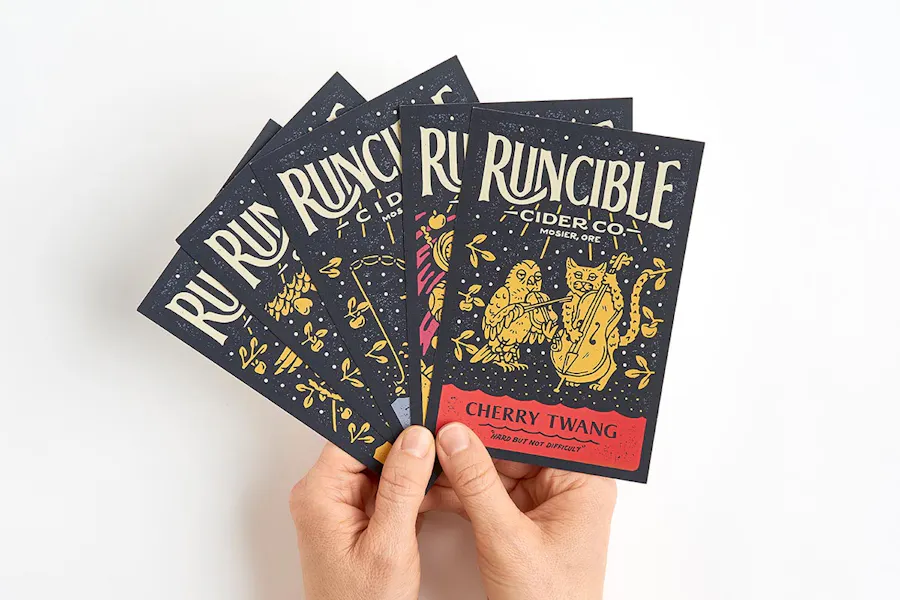 Two hands holding five fanned-out custom postcards printed with Runcible Cider and an owl and cat design.