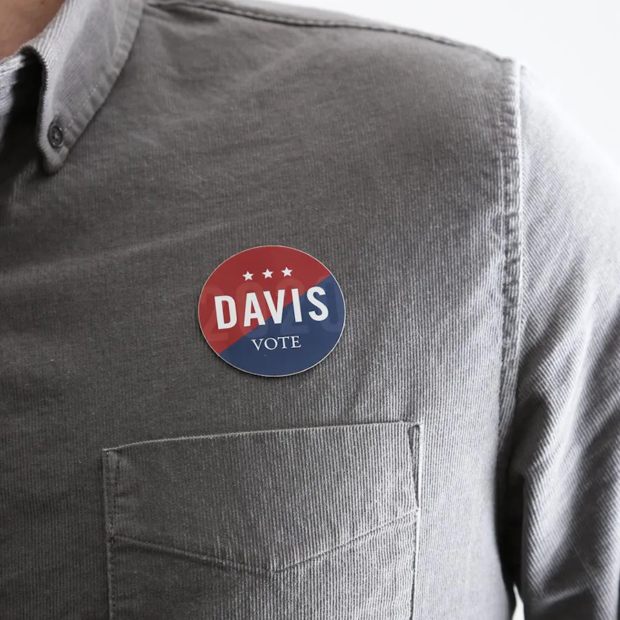 A person wearing a gray button-up shirt with a political campaign sticker above the chest pocket.