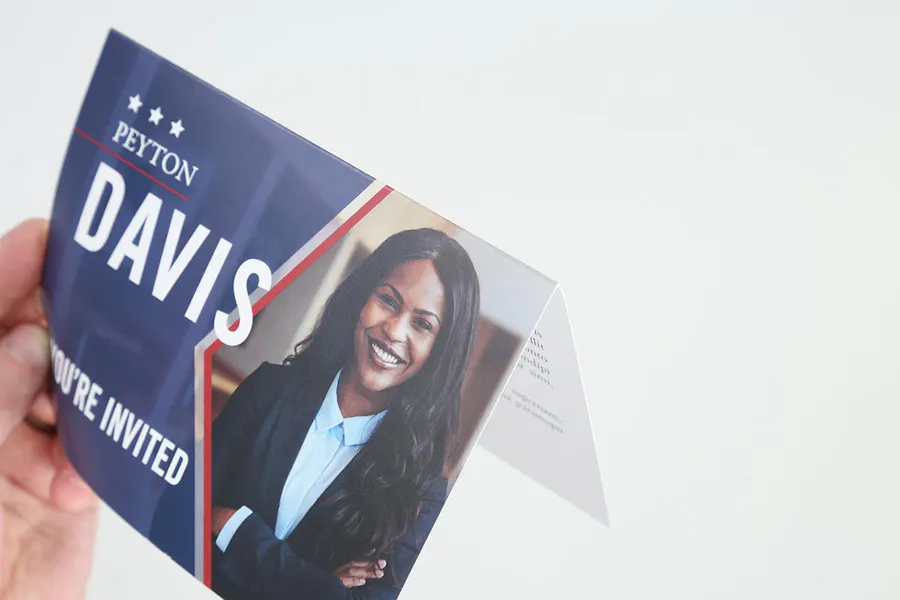 A hand holding a folded political mailer printed with a woman in a suit smiling on the front.
