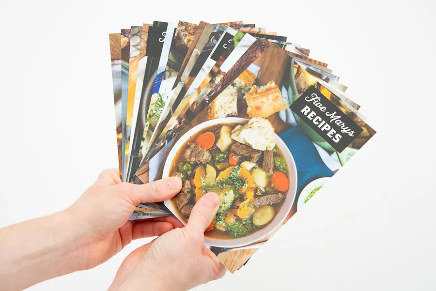 Two hands holding fanned-out recipe postcards with vibrant imagery on the front.