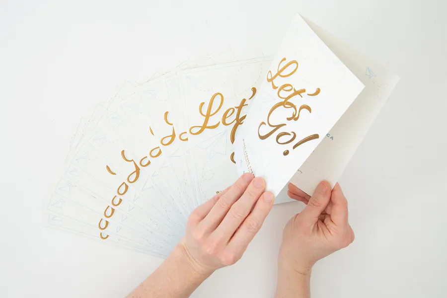 Two hands holding a personalized invitation with Let's Go! on the front and more fanned out invites below it.