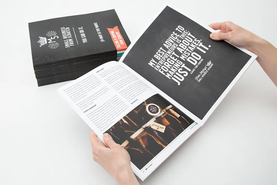 Two hands holding open a marketing booklet with a perfect binding in front of a stack of more booklets with black covers.