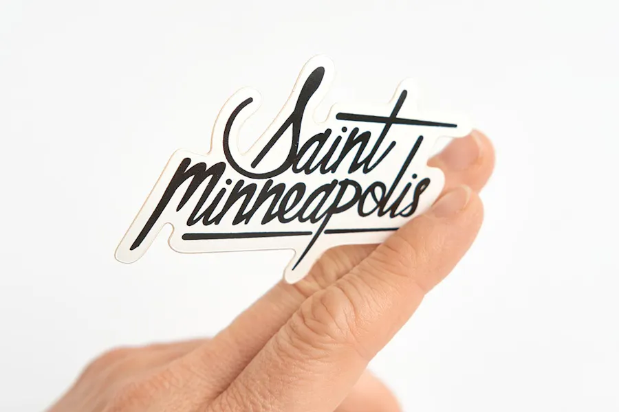 A hand holds a die-cut sticker between two fingers with Saint Minneapolis in black.