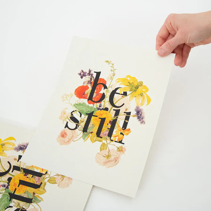 A hand taking a custom poster off a stack of posters printed with be still and a floral design.