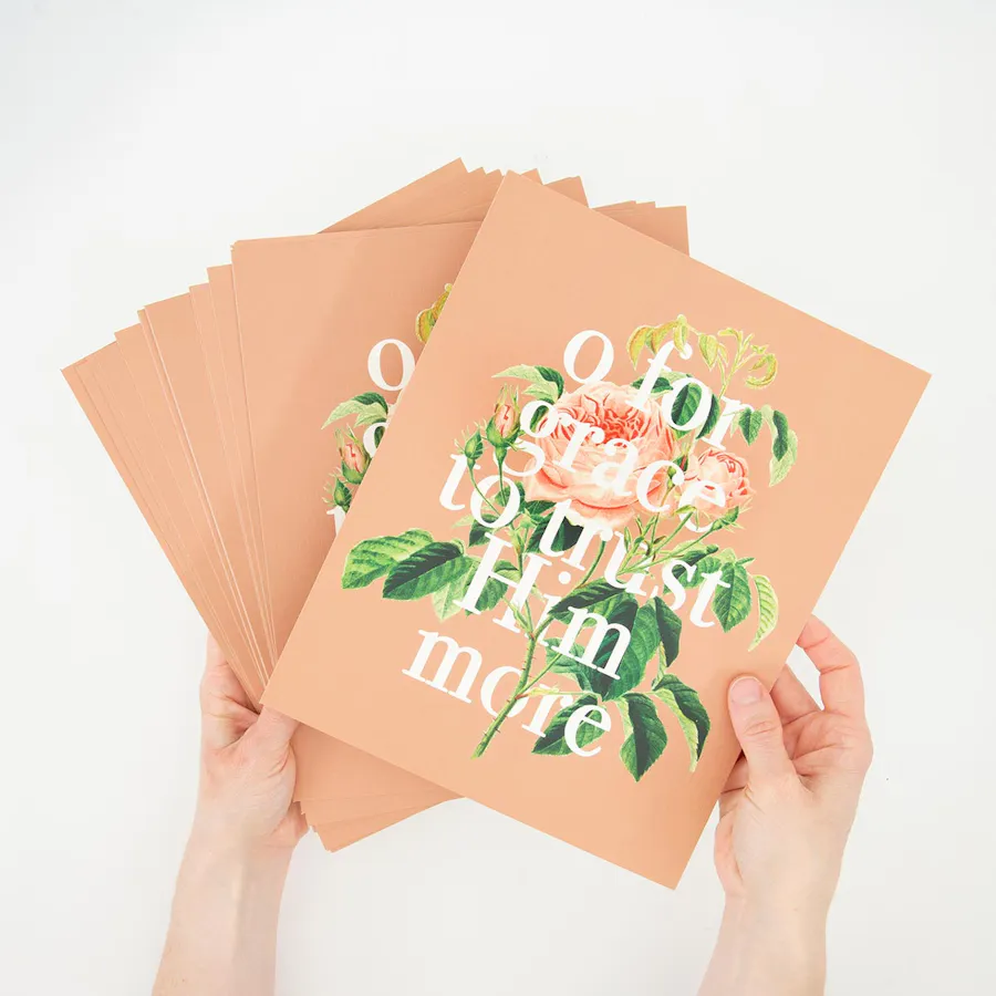 Two hands holding a stack of fanned-out custom posters with O for grace to trust Him more and a floral design.