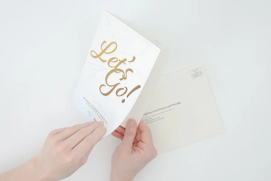 Two hands holding a nonprofit invite with Let's Go! on the front and a matching envelope laying underneath it.