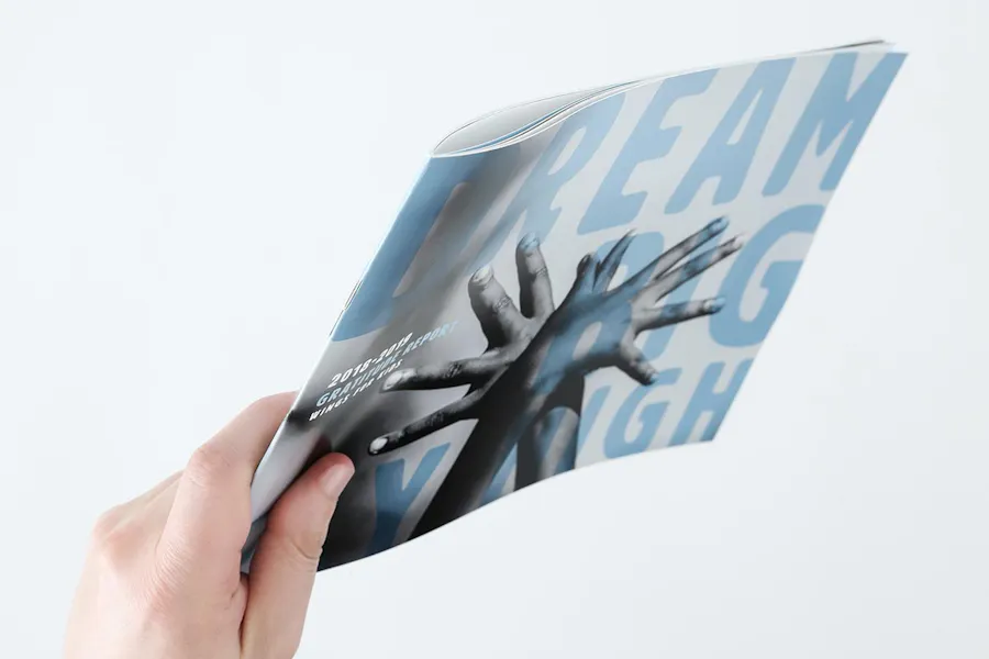 A hand holding a nonprofit annual report with Dream Big Fly High and two intertwined hands on the cover.