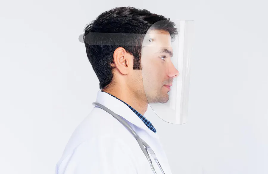A man with dark hair standing in profile wearing a lab coat, stethoscope and a clear protective face shield.