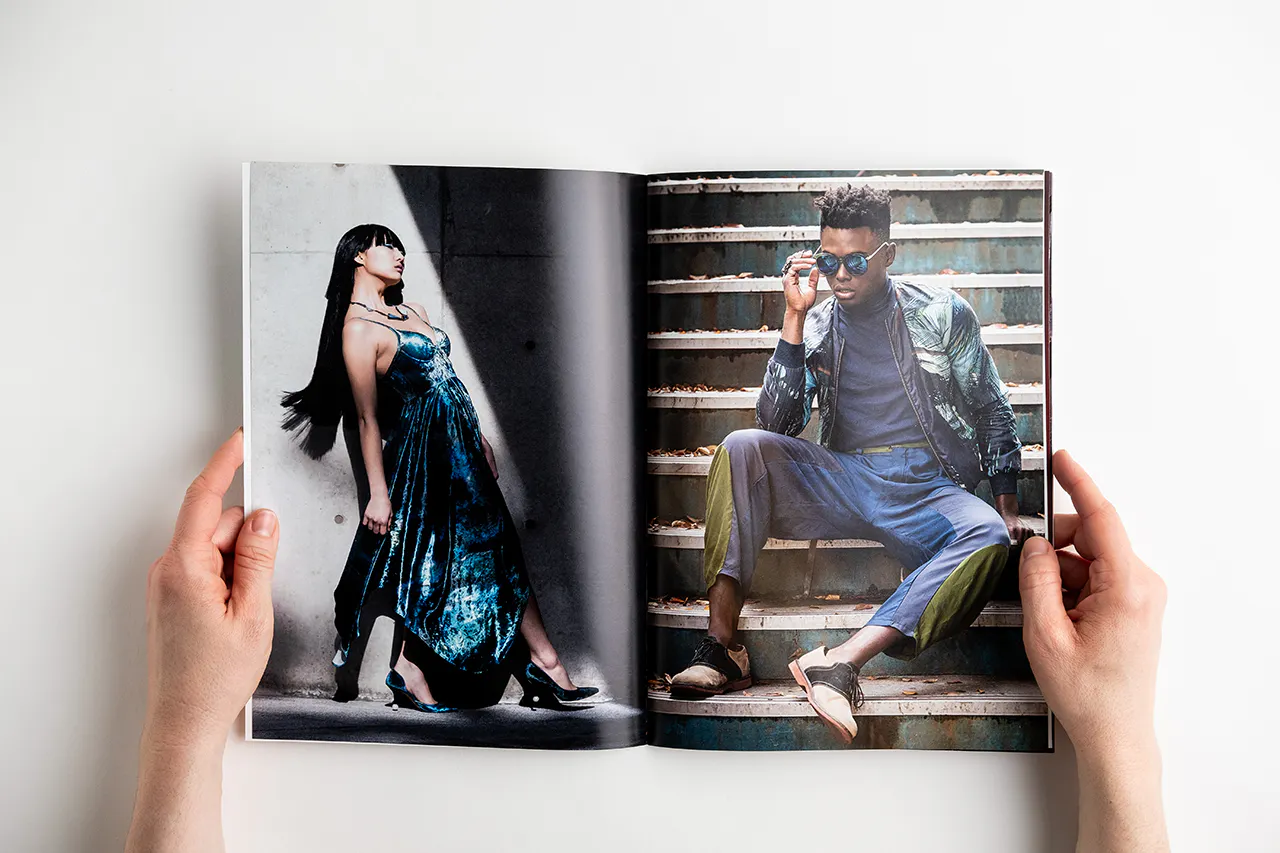 Two hands holding open a photography book with images of a woman in a dress and a man sitting on steps.