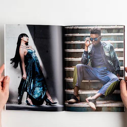 Two hands holding open a photography book with images of a woman in a dress and a man sitting on steps.