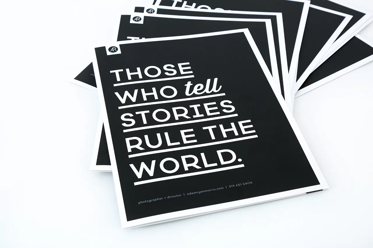 A stack of fanned-out photography booklets printed with a black cover and Those Who Tell Stories Rule the World in white.