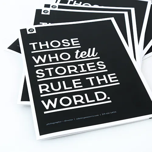 A stack of fanned-out photography booklets printed with a black cover and Those Who Tell Stories Rule the World in white.