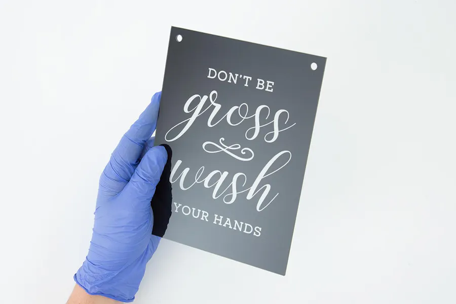 A gloved hand holding safety signage printed with Don't Be Gross Wash Your Hands in black and white.