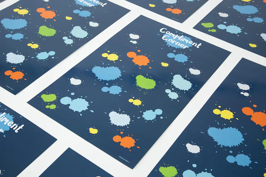 Custom classroom posters lined up in rows with a navy background, colorful splotches and Compliment Corner at the top.