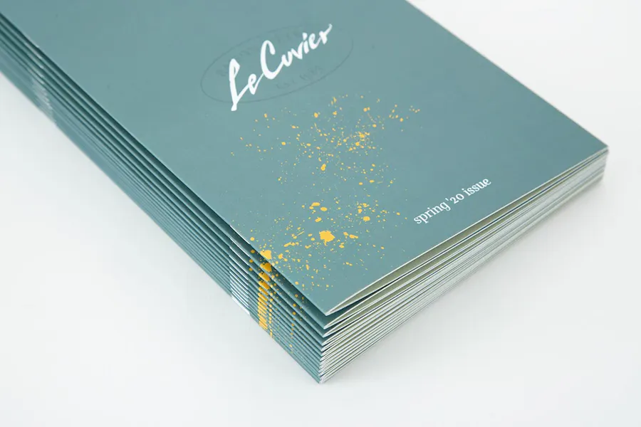 A stack of winery menus printed with a saddle stitch binding and a teal cover with "Le Cuvier" in white.