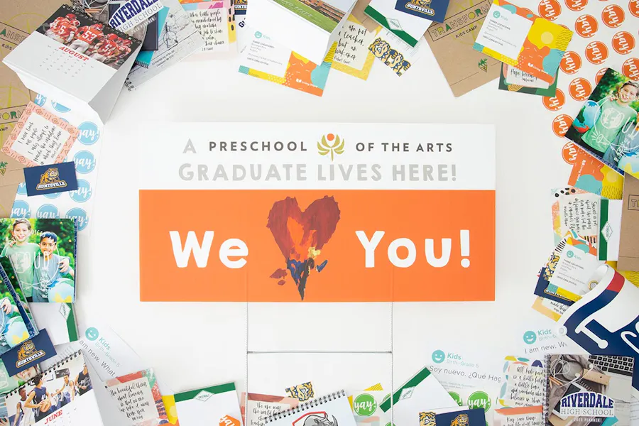 A graduation yard sign with a white and orange design surrounded by school print materials like planners and flashcards.