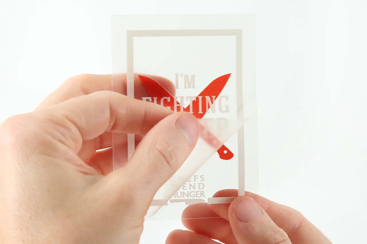 Two hands pulling back the liner on a clear decal printed with I'm Fighting over two red knives.