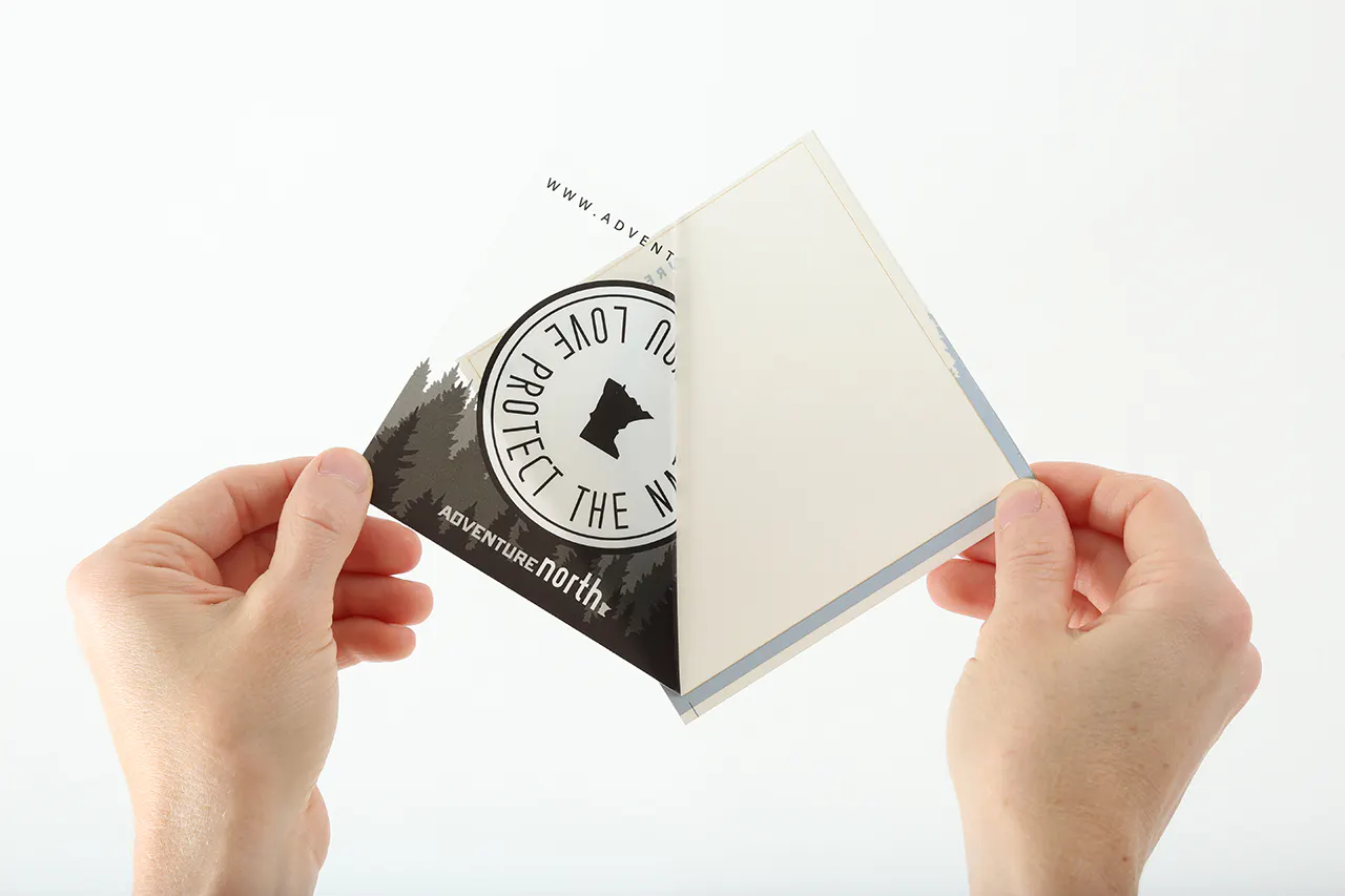 Two hands pulling off the liner of a custom sticker printed with Adventure North and a woods design.