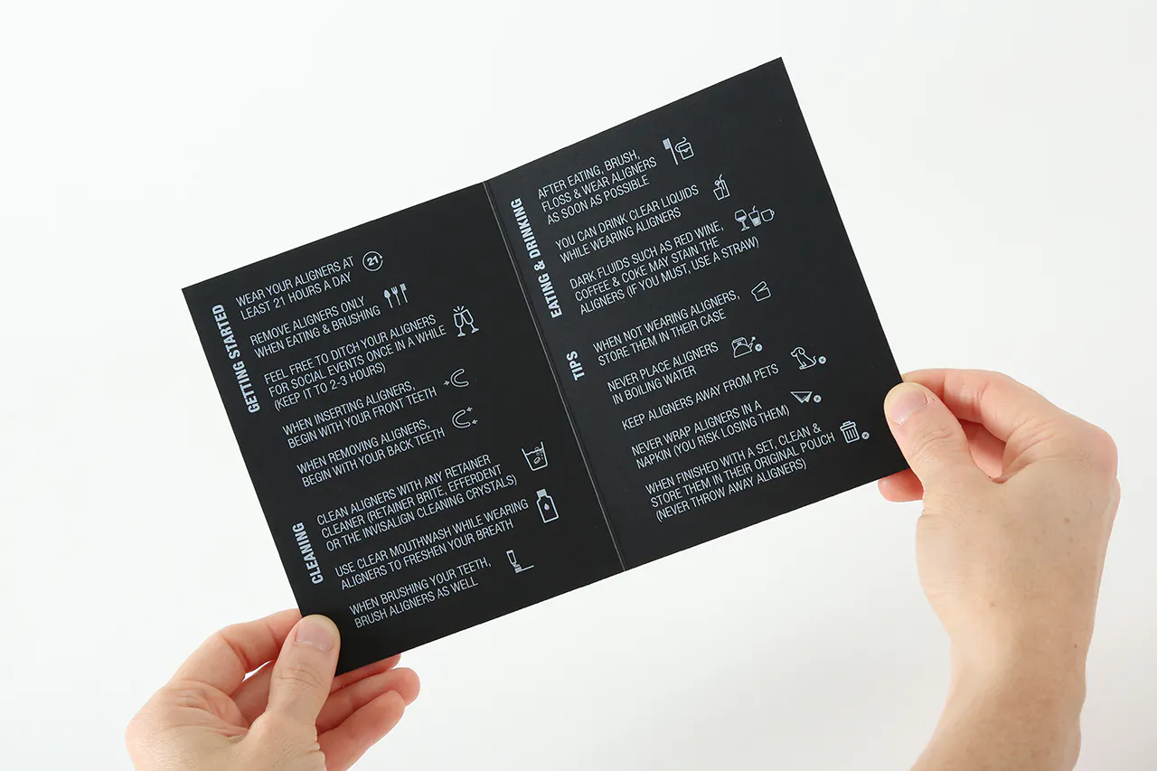 Two hands holding open care instructions for teeth aligners printed on black paper with white ink.
