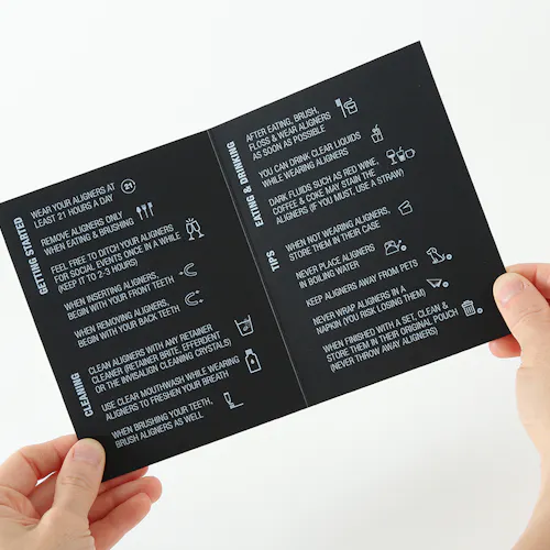Two hands holding open care instructions for teeth aligners printed on black paper with white ink.