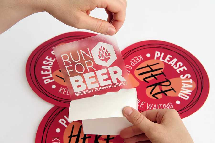 Two hands peeling a decal with Run for Beer in white letters away from its backing with red floor graphics underneath it.