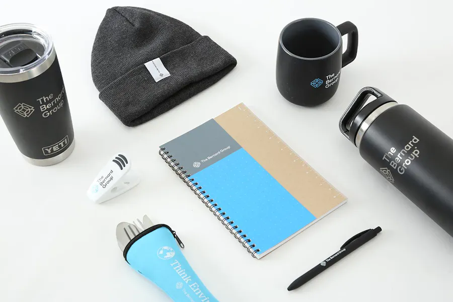 A promotional hat, notebook, pen, chip clip, silverware and mugs printed with The Bernard Group and its logo.