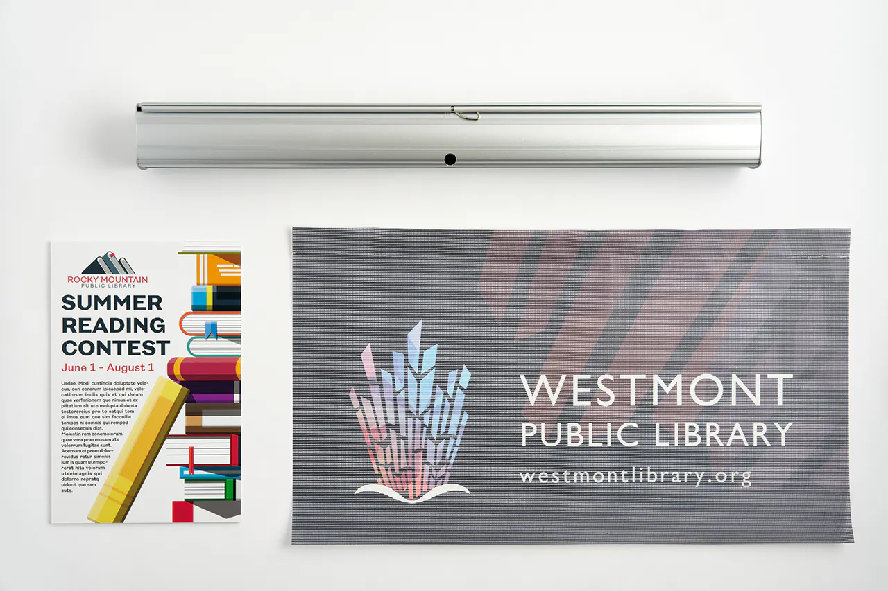 A retractable banner stand laying above a banner printed with Westmont Public Library and a book poster next to it.