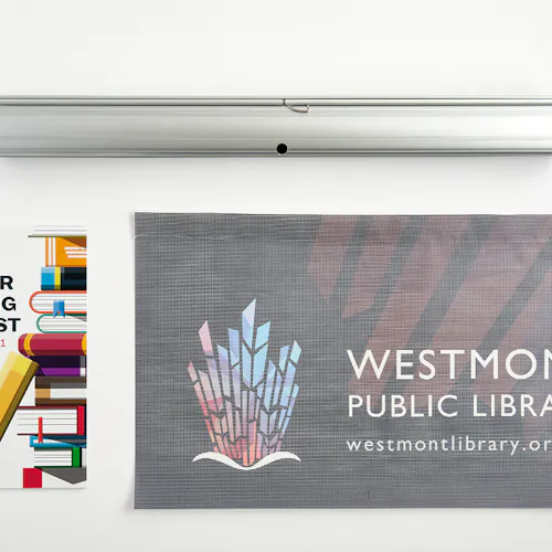 A retractable banner stand laying above a banner printed with Westmont Public Library and a book poster next to it.